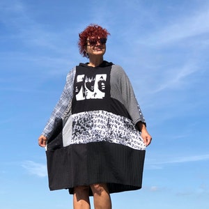 May include: A black and white patchwork dress with a hooded neckline. The dress features a graphic print of three faces and text that says "Dance Power" and "Sound" on the front. The dress is worn by a person with red hair and sunglasses.
