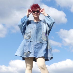 Op de afbeelding: Een vrouw draagt een blauwe denim jurk met patches en een hartvormig ontwerp. Ze draagt een zonnebril en heeft rood haar. De achtergrond is een blauwe lucht met witte wolken.