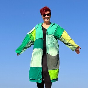 Könnte beinhalten: Eine Frau trägt einen langen Patchwork-Mantel aus verschiedenen Grüntönen und Gelb. Der Mantel hat eine Kapuze und ist vorne offen, wodurch ein schwarzes Shirt darunter sichtbar wird. Die Frau lächelt und hat rote Haare. Der Hintergrund ist ein klarer blauer Himmel.