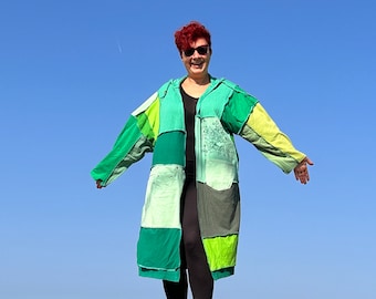 May include: A woman wearing a long, patchwork coat made of different shades of green and yellow fabric. The coat has a hood and is open in the front, revealing a black shirt underneath. The woman is smiling and has red hair. The background is a clear blue sky.