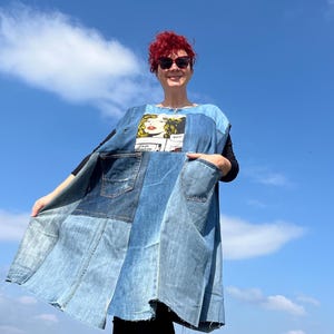 Puede incluir: Una mujer lleva un chaleco vaquero con un estampado de personaje de cómic y una camiseta negra. El chaleco está hecho de diferentes piezas de mezclilla cosidas juntas y tiene bolsillos en la parte delantera. La mujer está de pie frente a un cielo azul con nubes blancas.