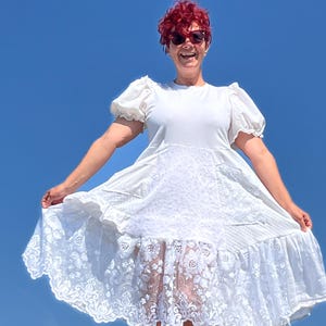 Könnte beinhalten: Ein weißes Kleid mit kurzen, gepufften Ärmeln und einem Spitzenrock. Das Kleid besteht aus einem weichen, fließenden Stoff und hat ein florales Spitzenmuster am Saum. Das Kleid ist knielang und wird anscheinend im Freien vor blauem Himmel getragen.