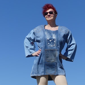 Puede incluir: Una mujer con un vestido de camisa vaquero azul con un bolsillo estampado con estrellas. El vestido tiene varios bolsillos y un corte holgado. Está de pie con las manos en las caderas y mirando hacia arriba.