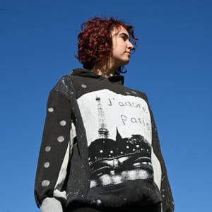 May include: A black hooded sweatshirt featuring a large white rectangular graphic on the front displaying the Eiffel Tower, a bridge, and the text J'adore Paris. The sleeves have light grey panels with white circular spots and grey cuffs.