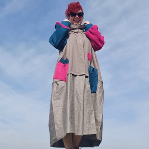 Könnte beinhalten: Eine Frau trägt ein langes, graues Leinenkleid mit bunten Patchwork-Details. Das Kleid hat große Taschen und einen Knopfleisten vorne. Die Frau trägt eine Sonnenbrille und hat rote Haare.