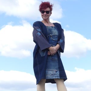 May include: A woman wearing a blue denim jacket with stars and a denim skirt. She is standing in front of a blue sky with white clouds.