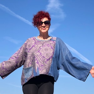 Puede incluir: Una mujer con un top de patchwork morado y azul con un patrón de paisley. El top tiene un bolsillo de mezclilla en la parte delantera. Lleva pantalones negros y tiene el pelo rojo rizado.