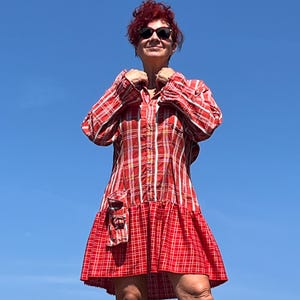Op de afbeelding: Een rood en wit geruit overhemdjurk met lange mouwen en een geplooid zoom. De jurk heeft een zak aan de voorkant en wordt buiten gedragen tegen een heldere blauwe lucht. De jurk heeft een knoopsluiting.