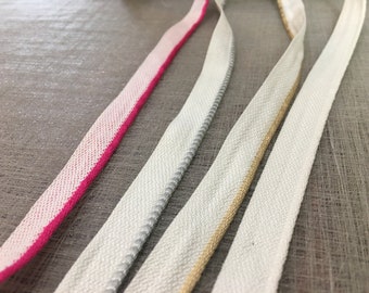 May include: Four white fabric trims with different colored edges. The trims are pink, silver, gold, and white.