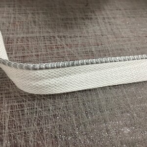 May include: White fabric trim with a silver edge. The trim is on a textured surface.
