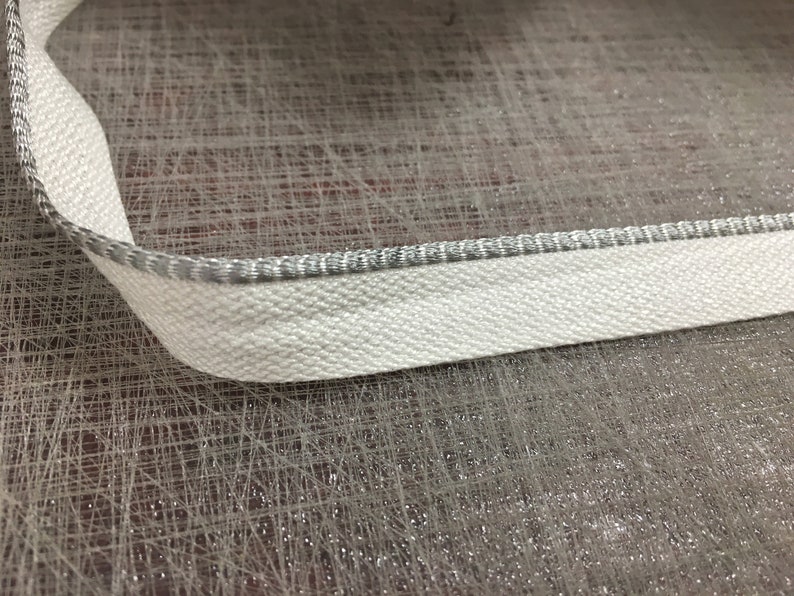May include: White fabric trim with a silver edge. The trim is on a textured surface.