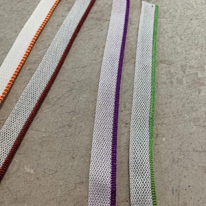 May include: Five different white fabric trims with colored stripes. The trims have a woven texture and are arranged vertically.