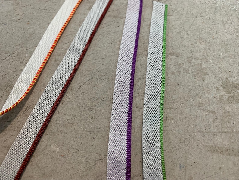 May include: Five different white fabric trims with colored stripes. The trims have a woven texture and are arranged vertically.