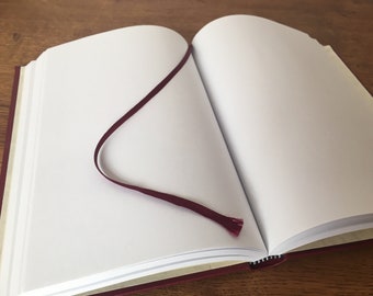 May include: A burgundy notebook with a blank page open and a burgundy ribbon bookmark. The notebook is lying on a wooden surface.
