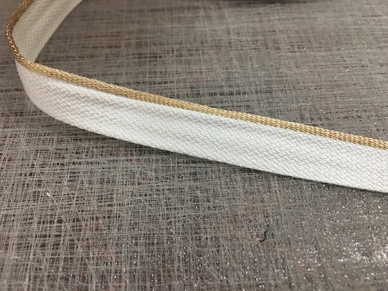 May include: White cotton ribbon with a gold trim. The ribbon is flat and has a woven texture.