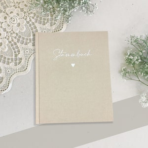 Può includere: Un diario beige in tessuto di lino con la parola "Stammbuch" scritta in corsivo bianco e un piccolo cuore bianco.
