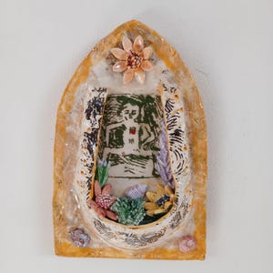 Puede incluir: Un adorno de pared de cerámica con forma de barco. La pieza presenta una imagen central de una persona, rodeada de elementos florales y conchas marinas. La paleta de colores incluye tonos beige, verde, rosa y amarillo, con detalles decorativos en negro.