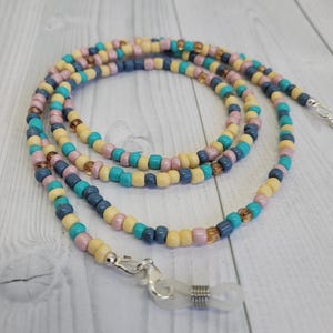 Könnte beinhalten: Eine Brillenbandkette aus Perlen in einem Spiralmuster. Die Kette besteht aus kleinen, farbigen Perlen in Türkis, Gelb, Rosa, Marineblau und Braun. Sie hat silberfarbene Verschlüsse zum Befestigen an Brillen.