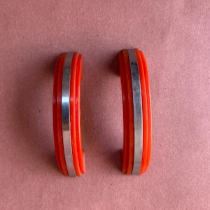 Puede incluir: Dos tiradores de cajón semicirculares con un acabado naranja vibrante y una incrustación central vertical de metal plateado. Cada tirador presenta un diseño acanalado y escalonado en los bordes exteriores.