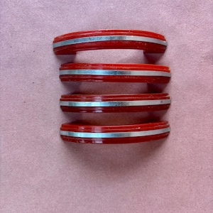 May include: Four matching drawer pulls arranged in a vertical stack. Each pull features a curved, bright red body with a smooth, glossy finish and a single, centred horizontal silver-toned metal inlay running across the entire length of the handle.