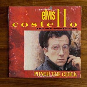 May include: Album cover for "Punch the Clock" by Elvis Costello and The Attractions. The cover features a portrait of a man wearing glasses and a hat, set against a red background. The album title and artist's name are in yellow text.