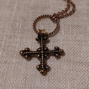Puede incluir: Un collar de cadena dorada con un colgante de cruz de bronce adornado y detallado. La cruz tiene un diseño de cuerda y está adornada con pequeños acentos redondos.
