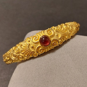 May include: A gold-toned bangle bracelet with an intricate, ornate design. The bracelet features a single red gemstone in the center.