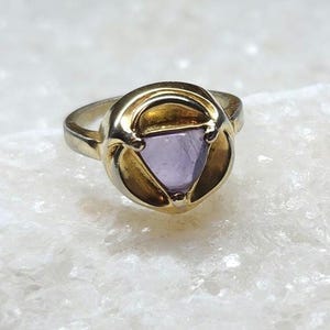 May include: A gold-toned ring with a triangular, light purple gemstone set in a decorative bezel. The ring's band is a simple, polished gold. The ring is set against a white, textured surface.