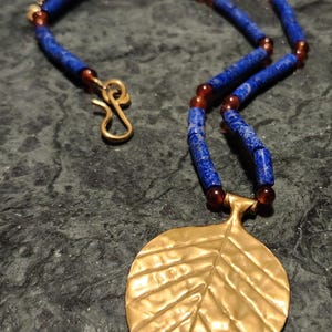 May include: A necklace featuring a gold-colored leaf pendant and a beaded chain. The chain is composed of blue and brown beads, with a gold-colored clasp. The leaf pendant has a textured surface and is the focal point of the necklace.