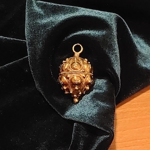 May include: A gold-colored, ornate, spherical pendant with a textured surface and a pointed bottom. The pendant is hanging from a loop at the top.