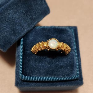 May include: A gold ring with an opal gemstone, set in a textured band. The ring is displayed in a dark blue velvet ring box. The opal is round and has a milky white appearance.