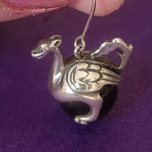 May include: A silver-toned earring featuring a sculpted dragon figure with detailed wings and a long neck, positioned over a smooth, spherical black bead. The dragon is attached to a thin metal hook for suspension, creating a dangling design.