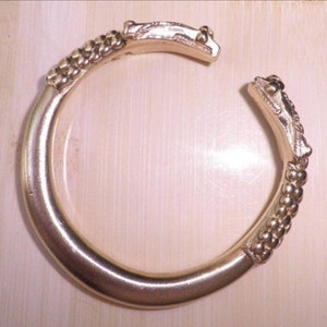 May include: A gold-toned bangle bracelet with a textured surface and a decorative design on the ends. The ends of the bracelet are shaped like a dragon's head.