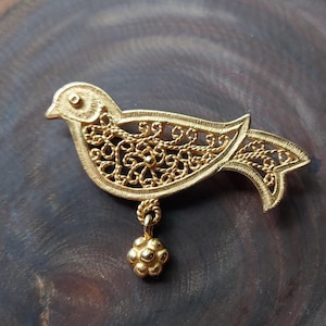 Puede incluir: Un broche de metal dorado con forma de pájaro, con un contorno texturizado y un intrincado diseño de ala en filigrana. Un pequeño dije redondo en forma de flor cuelga de la parte inferior del cuerpo del pájaro mediante un aro de metal trenzado.