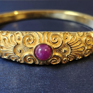 May include: A gold-colored bangle bracelet with intricate floral and scrollwork designs. The bracelet features a central, round, red gemstone. The jewelry is displayed on a dark blue surface.