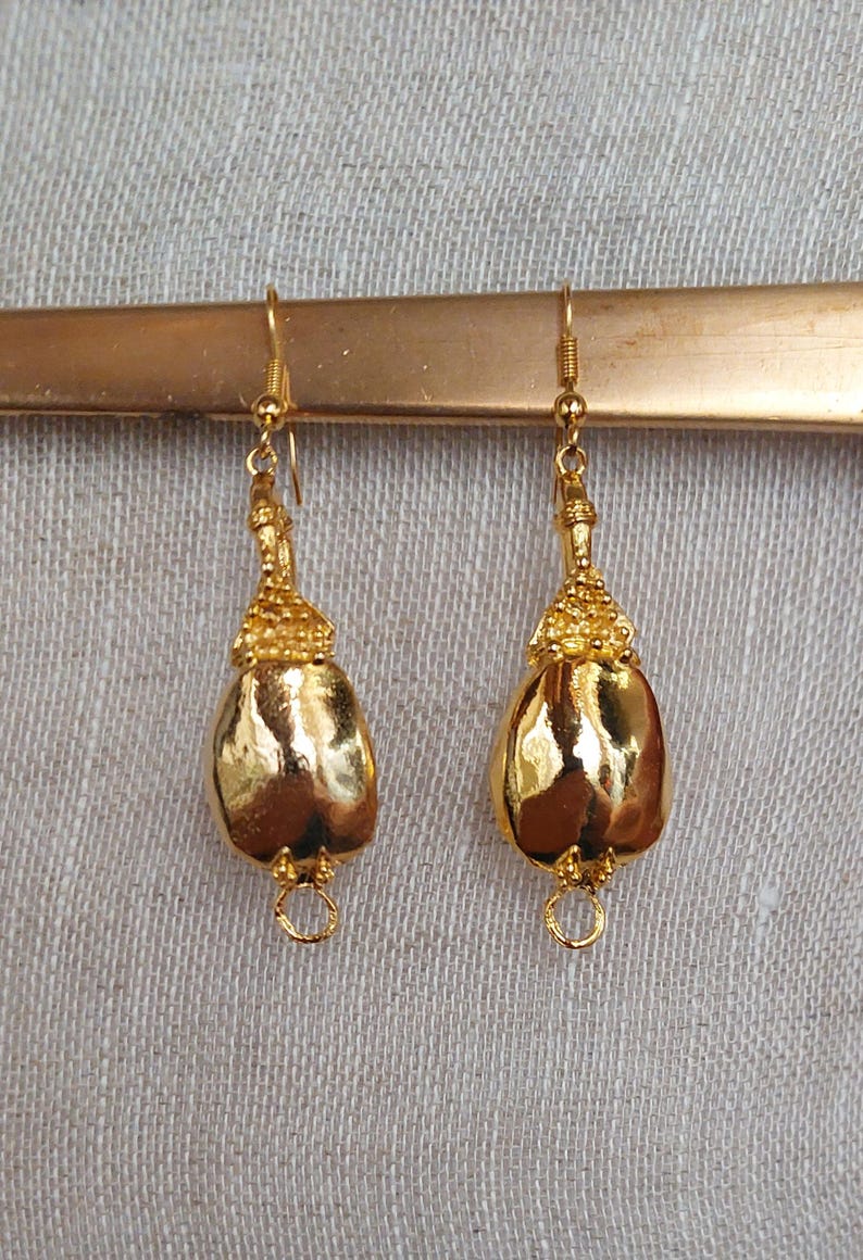 Puede incluir: Un par de pendientes dorados con forma de l&aacute;grima. Los pendientes tienen una parte superior texturizada y un cuerpo liso y brillante. Cuelgan de un gancho dorado y tienen un peque&ntilde;o anillo en la parte inferior.