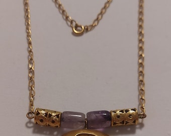 May include: A gold chain necklace with a large, oval pendant featuring a cut-out circle. The pendant is flanked by two amethyst beads and two gold beads with intricate designs.