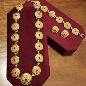 May include: A gold-toned jewelry set featuring a necklace, bracelet, and earrings. The pieces have a circular design with a floral pattern and a small, dark red center. The set is displayed on a burgundy velvet surface.