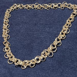 May include: A gold-colored necklace with a chainmail design. The necklace is composed of small, interconnected rings, creating a delicate and intricate pattern. The jewelry is displayed on a dark blue surface.