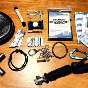 Puede incluir: Un conjunto de equipo de supervivencia que incluye una bolsa de cuero negro, una manta de rescate de lámina, una multiherramienta, una brújula y varias herramientas pequeñas y accesorios. Los artículos están dispuestos sobre una superficie de madera.