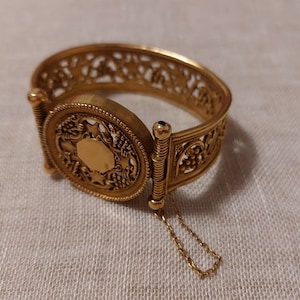 Puede incluir: Una pulsera dorada con bisagras, con una pieza central ovalada y un intrincado diseño calado. La pieza central presenta una forma ovalada en blanco rodeada por un patrón detallado de uvas y hojas. Se adjunta una delicada cadena.