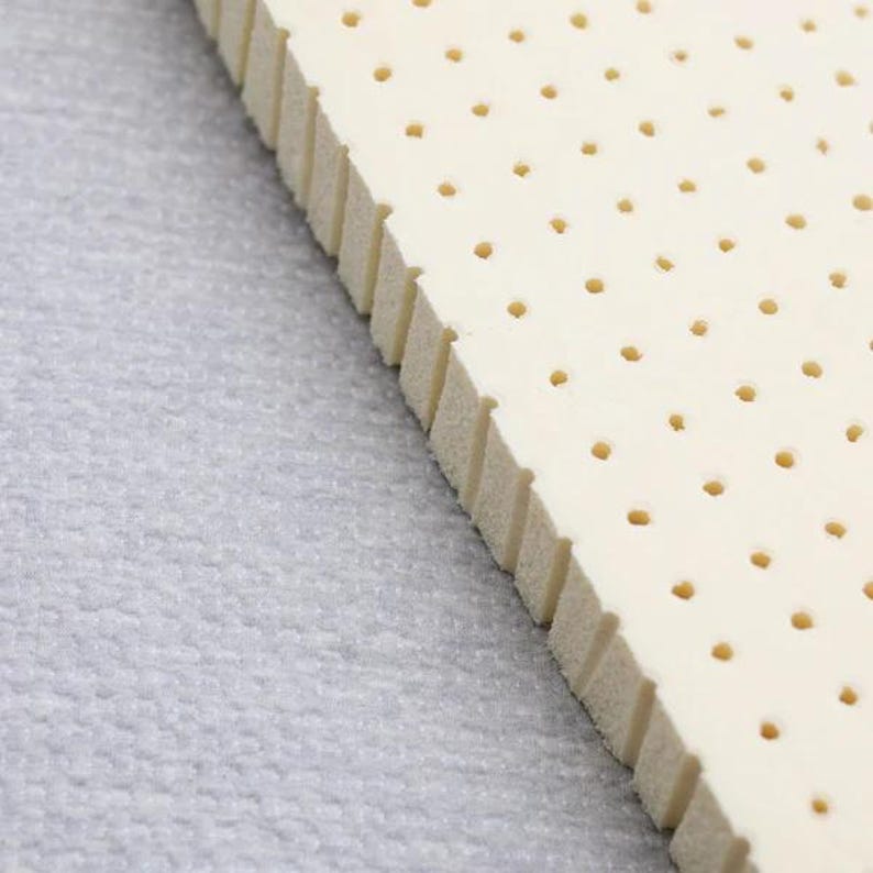 K&ouml;nnte beinhalten: Nahaufnahme eines wei&szlig;en Latex-Matratzenauflages mit kleinen L&ouml;chern &uuml;ber die gesamte Fl&auml;che. Das Auflage ist auf einer grauen Oberfl&auml;che.