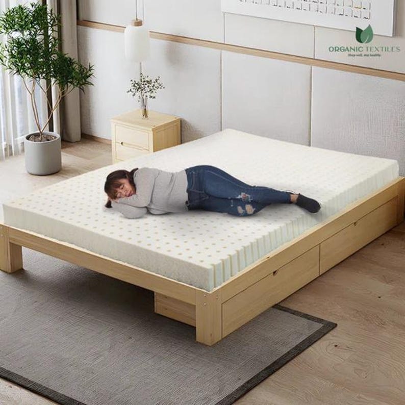 K&ouml;nnte beinhalten: Eine Person liegt auf einer wei&szlig;en Matratze mit einem Holzrahmen. Die Matratze hat ein Muster aus kleinen L&ouml;chern und ist aus organischen Materialien hergestellt. Der Rahmen hat zwei Schubladen an der Seite. Der Text "ORGANIC TEXTILES" ist in der oberen rechten Ecke des Bildes sichtbar.
