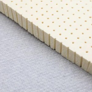K&ouml;nnte beinhalten: Nahaufnahme eines wei&szlig;en Latex-Matratzenauflages mit einem Raster aus kleinen L&ouml;chern. Der Topper ist schr&auml;g und die Kante ist sichtbar.
