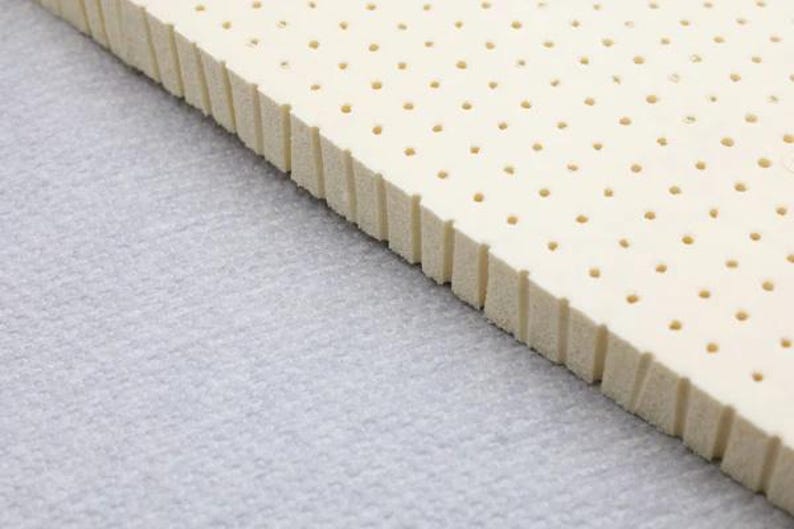 K&ouml;nnte beinhalten: Nahaufnahme eines wei&szlig;en Latex-Matratzenauflages mit einem Raster aus kleinen L&ouml;chern. Der Topper ist schr&auml;g und die Kante ist sichtbar.