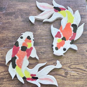 May include: Three ceramic koi fish sculptures with white, red, yellow, orange, and black markings. The fish are arranged on a wooden surface, showcasing their detailed design and vibrant colors. These decorative pieces are ideal for home decor.