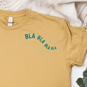 Può includere: T-shirt giallo senape con maniche arrotolate. La maglietta presenta la scritta "BLA BLA BLA" in verde acqua. La maglietta è appoggiata su una superficie bianca, con un cappello di paglia, una pianta succulenta e un cuscino decorativo sullo sfondo.