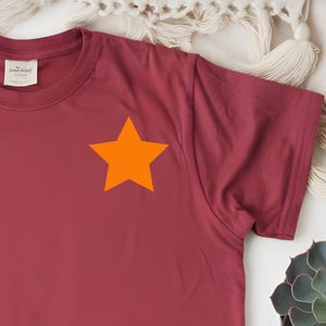 Könnte beinhalten: Ein weinrotes T-Shirt mit einem leuchtend orangefarbenen Stern auf der linken Brust. Das Shirt wird auf einer weißen Oberfläche mit dekorativen Kissen, einer Sukkulentenpflanze und einem Strohhut präsentiert.
