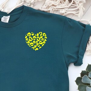 Può includere: Una t-shirt a maniche corte color verde acqua scuro con una piccola grafica a forma di cuore sulla parte superiore sinistra del petto. Il design del cuore presenta un motivo leopardato giallo neon brillante sul tessuto scuro.