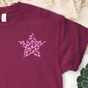 Può includere: T-shirt bordeaux con maniche arrotolate. Una stella rosa con motivo leopardato è sul petto sinistro. La maglietta è realizzata in materiale morbido, adatta per l'abbigliamento casual.